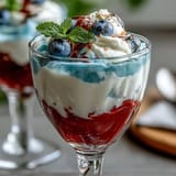 Festive red, white, and blue Jello cups in clear glasses, topped with whipped cream and fresh berries for a fun dessert.