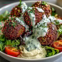 Fresh, crispy falafel balls rest atop creamy hummus and bright greens in a vibrant Falafel Bowl, drizzled with tahini sauce. 